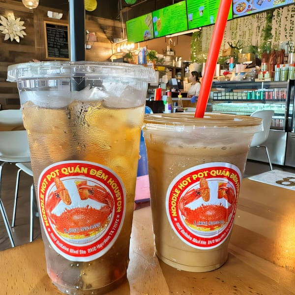 Two drinks with Quán Gì Quán Đêm Houston crab logo stickers on a wooden table inside the restaurant.