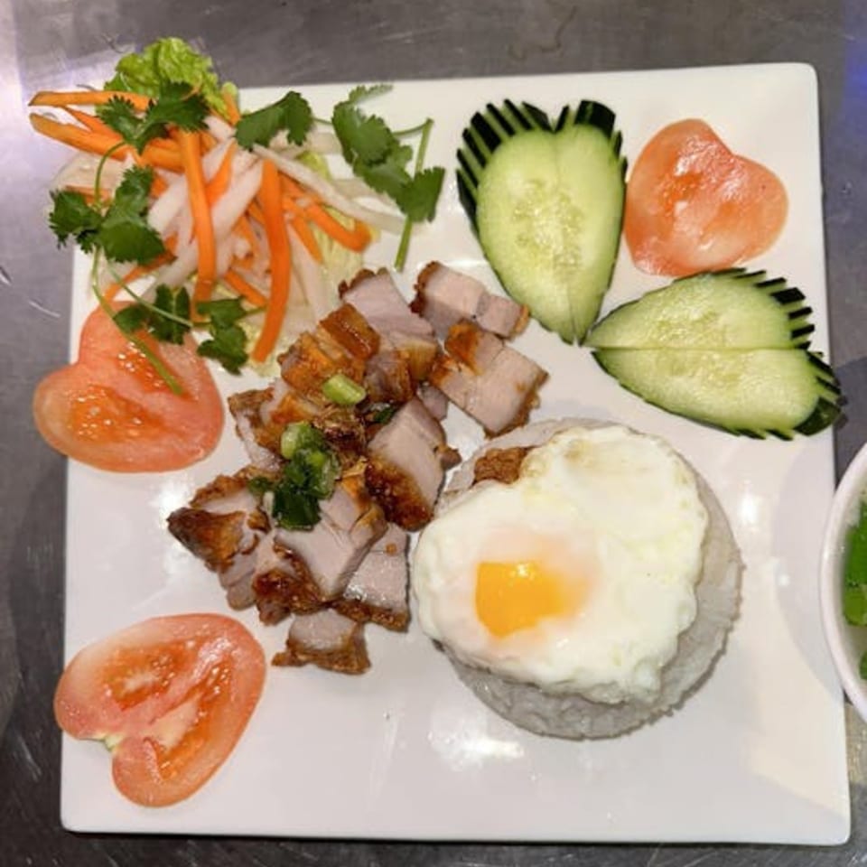 Vietnamese crispy pork belly with rice, a fried egg, pickled vegetables, and heart-shaped tomatoes and cucumbers.