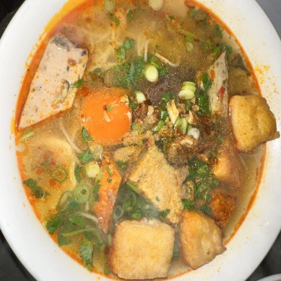 A close-up of a vibrant Vietnamese noodle soup with tofu, meat, and fresh herbs in a white bowl.