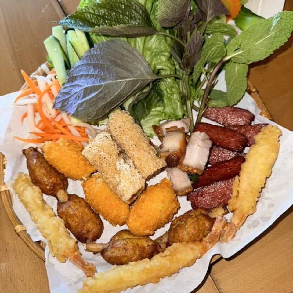 A Vietnamese platter with fried shrimp, sausage, pork belly, vegetables, and herbs on a woven tray.