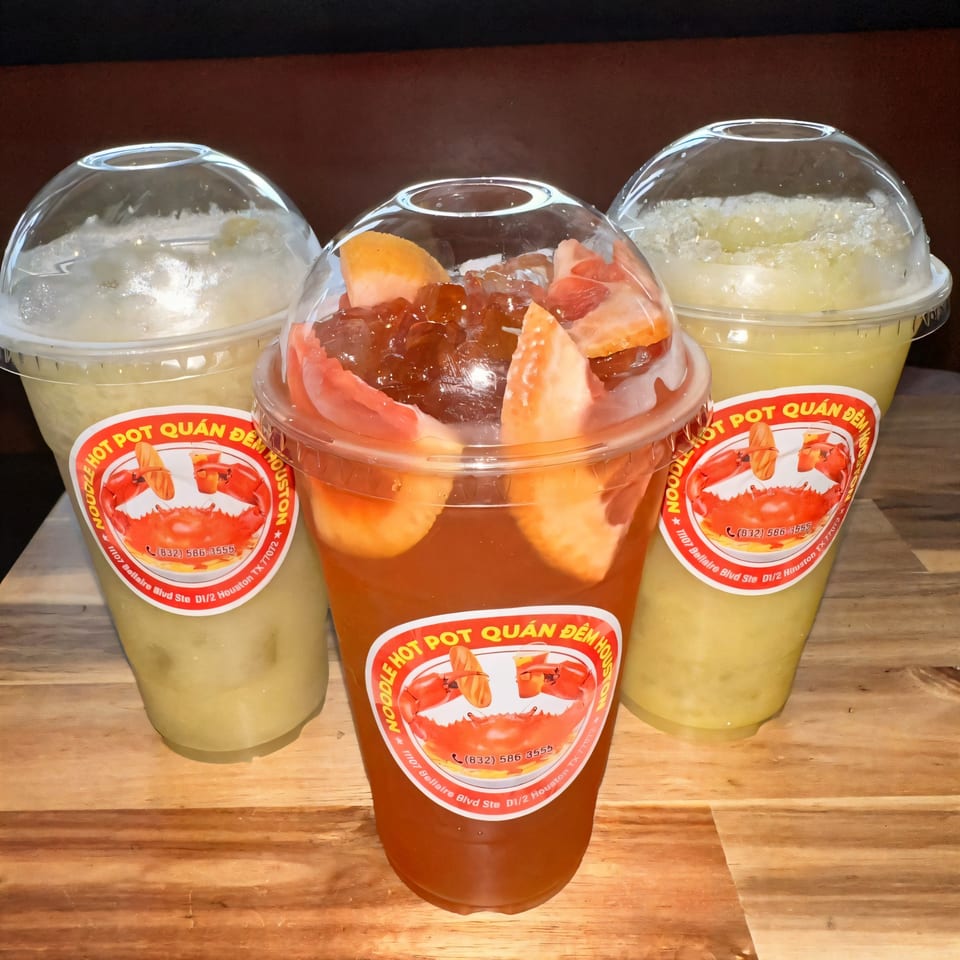 Three Quán Gì Quán Đêm Houston drinks, two light yellow and one red with fruit, on a wooden surface.
