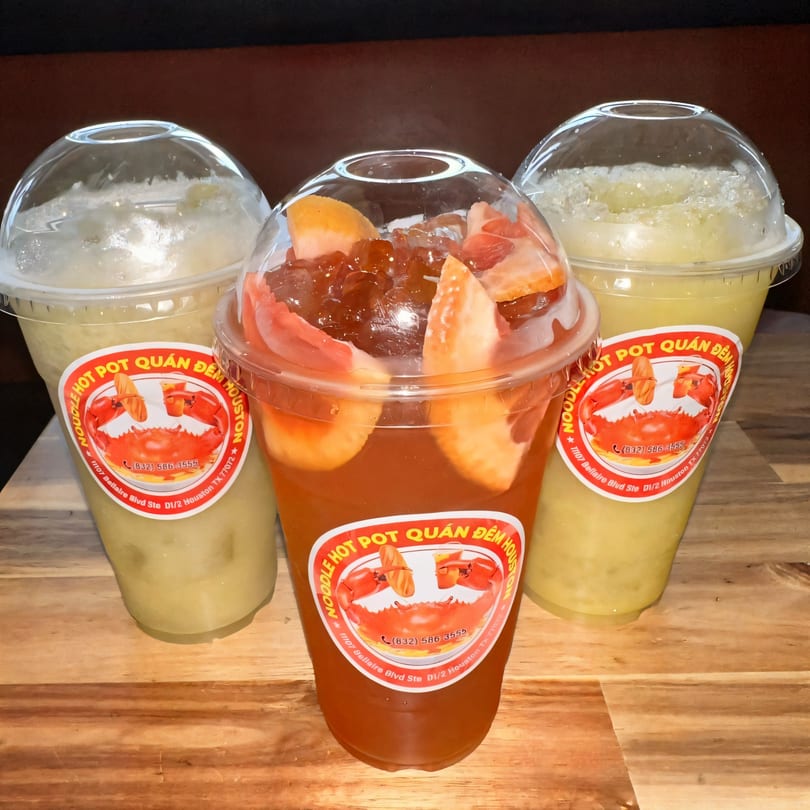 Three Quán Gì Quán Đêm Houston drinks, two light yellow and one red with fruit, on a wooden surface.